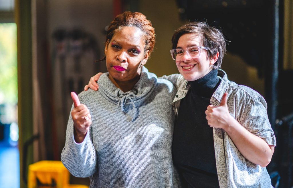 Two people stand together indoors, smiling and giving thumbs up. The atmosphere is warm and friendly, with soft lighting highlighting their faces.