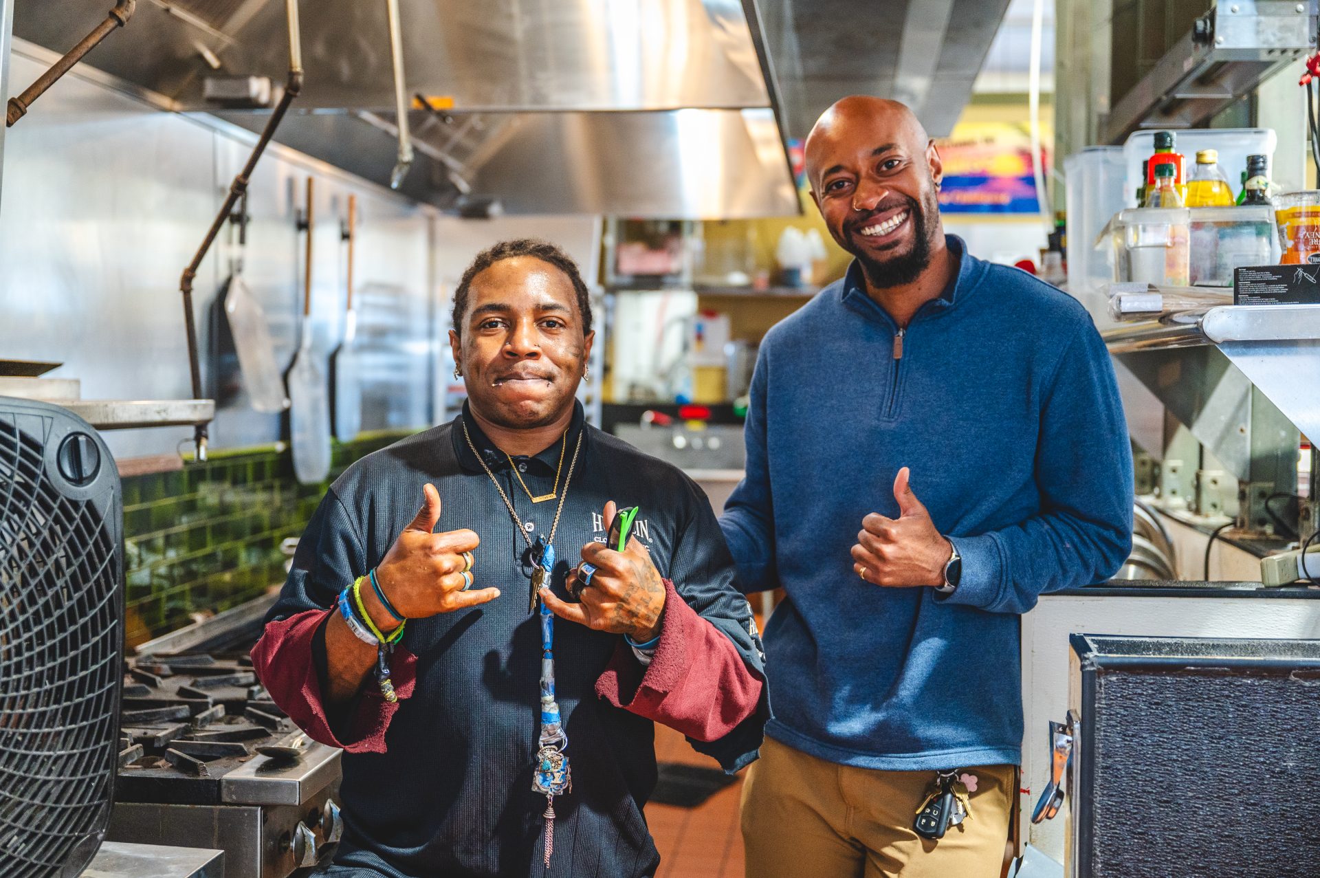Two Black men smiling in a restaurant kitchen. One in a dark chef's outfit, the other in a blue top and beige pants. Each gives a thumbs-up sign.