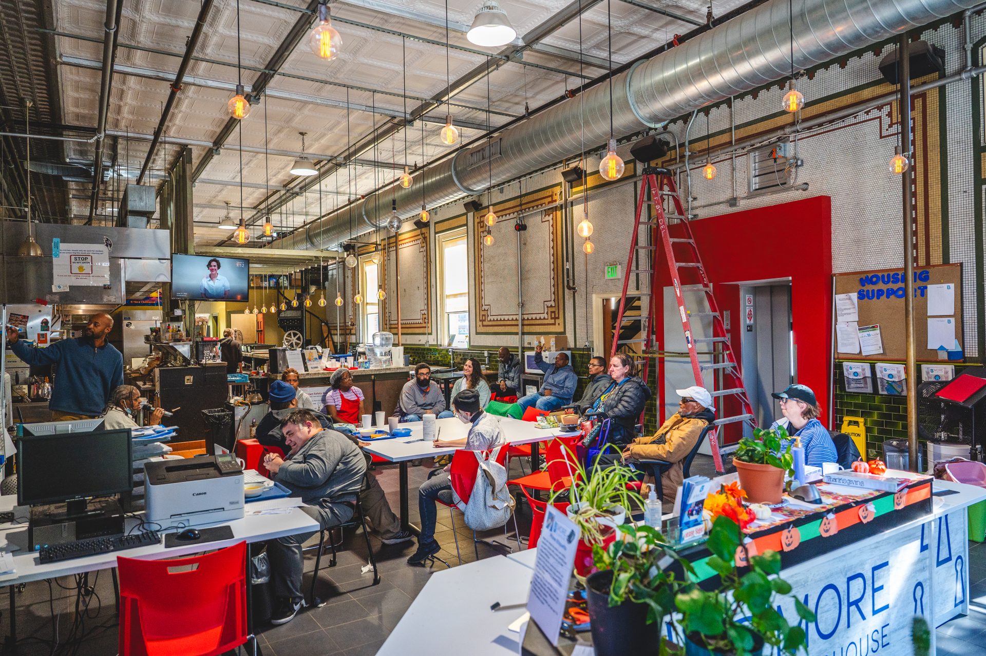 A diverse group of people sits at tables in a spacious, industrial-style room decorated with plants and warm lighting. A speaker addresses the group, conveying a collaborative and focused atmosphere.