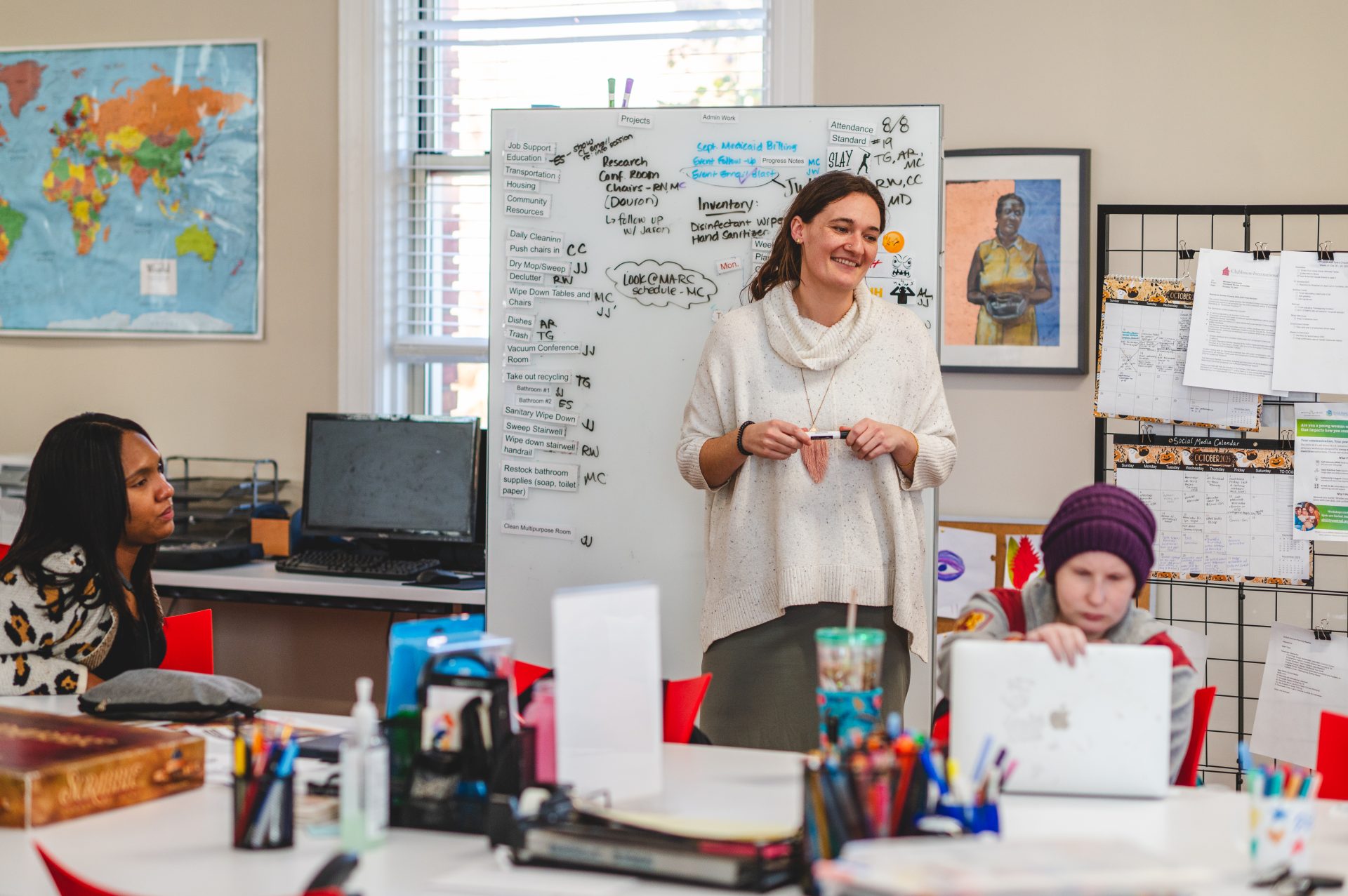 A woman smiles while standing near a whiteboard filled with notes in a classroom. Members focus on their laptops at a table. The room is colorful.