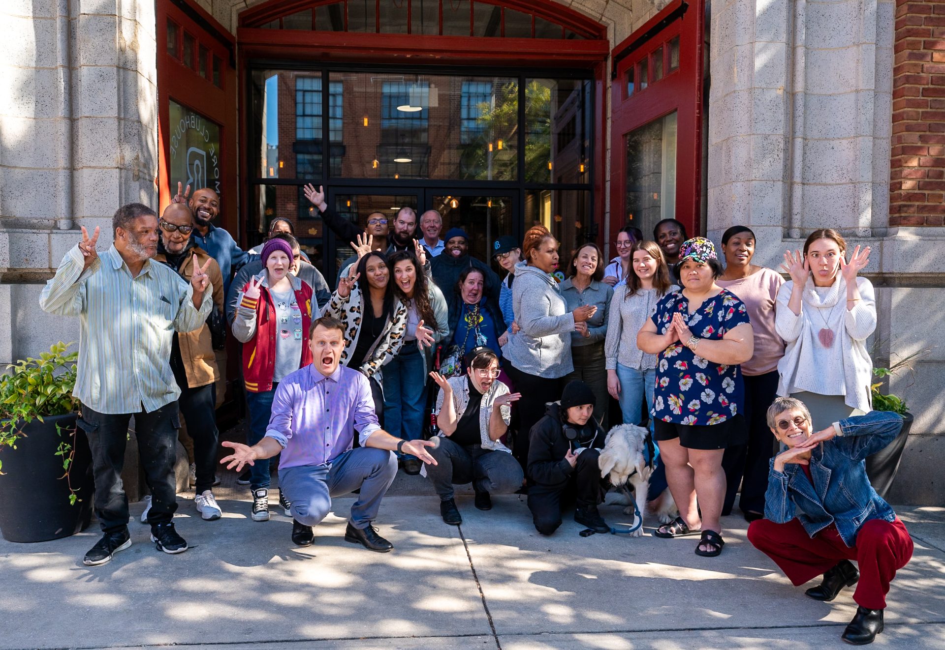 A diverse group of people joyfully posing outside B'More Clubhouse, a building with arched windows and red trim. They display playful gestures and smiles, creating a lively atmosphere.